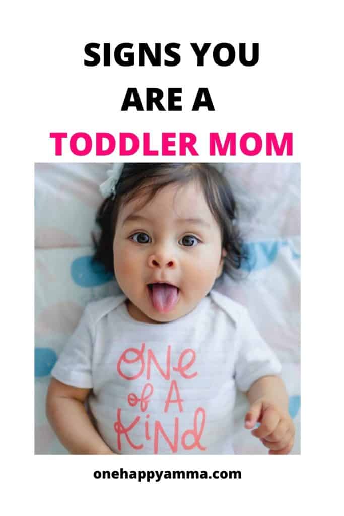 A toddler lying on a bed with her tongue out wearing one of a kind onesie used as a pinterest poster for Signs you are a toddler mom.